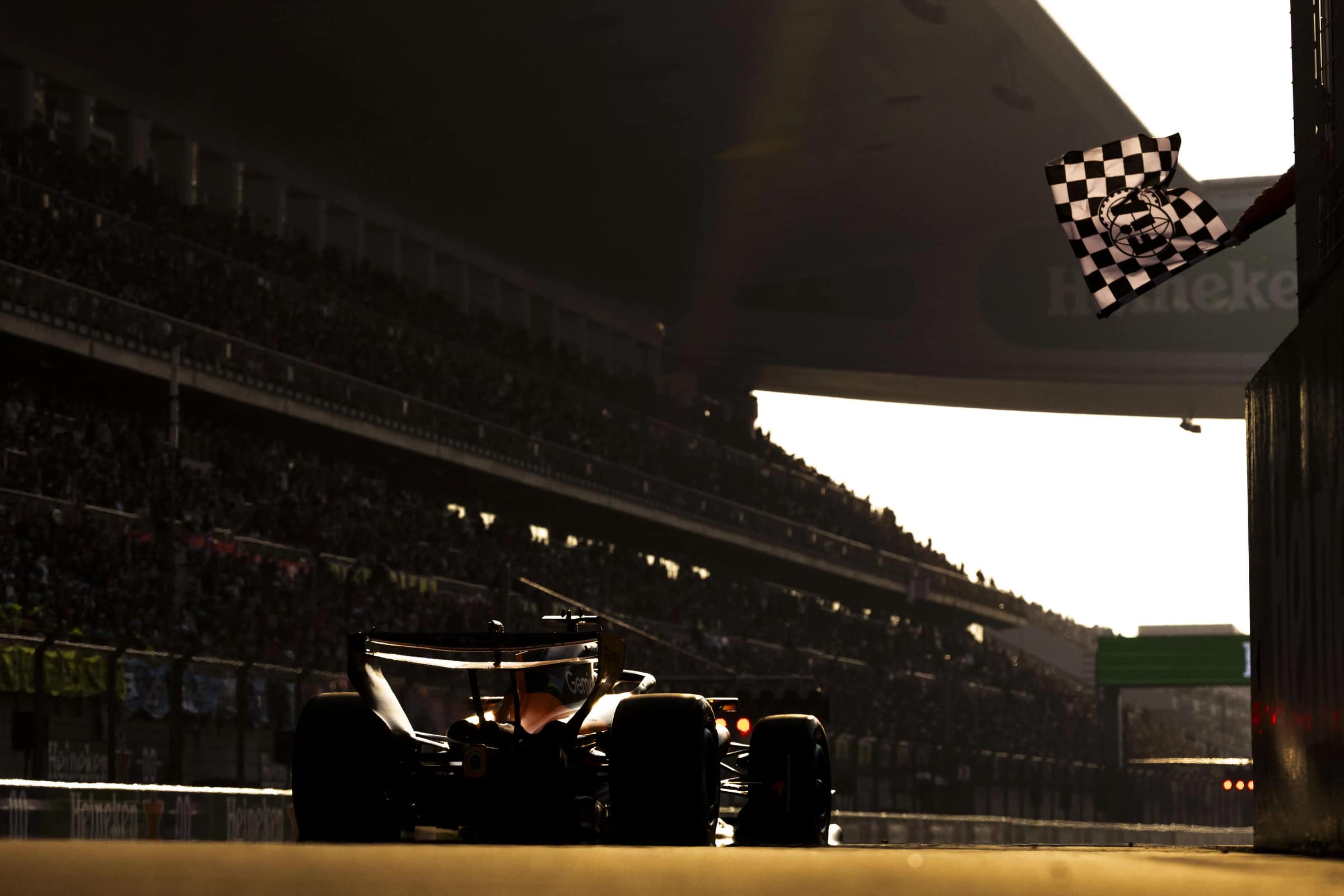 A racing car crossing the finish line under a chequered flag, the grandstand in silhouette
